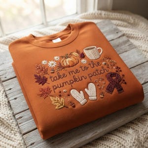 May include: A burnt orange crewneck sweatshirt featuring embroidered autumn-themed designs including a pumpkin, a mug, mittens, a scarf, and fall leaves. The text "take me to the pumpkin patch" is stitched in the centre in a dark brown script font.