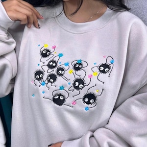 May include: A pale grey sweatshirt with an embroidered design of black, round creatures with white eyes and colourful accents. The design includes blue, yellow, and pink embroidered details. The sweatshirt has long sleeves.