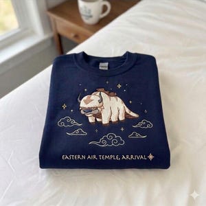 Może przedstawiać: Granatowa bluza z haftowanym wzorem Appa z Avatar: The Last Airbender. Wzór zawiera Appa, chmury, gwiazdy i tekst "Eastern Air Temple, Arrival". Bluza jest złożona na białej powierzchni.
