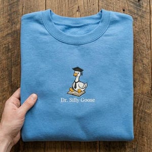 May include: A light blue crewneck sweatshirt featuring an embroidered graphic of a white goose wearing a black graduation cap and a black necktie while sitting on an open book. Below the goose, the text Dr. Silly Goose is embroidered in white.