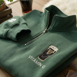 Pode incluir: Camisola verde com fecho de correr de um quarto, fecho de correr prateado e design de copo Guinness bordado. A camisola apresenta o texto "VITAMIN C" e um trevo branco no punho.