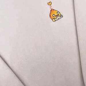 Op de afbeelding: Een lichtbeige sweatshirt met een klein geborduurd cartoonfiguur. Het figuur is oranje met een gele vlam en een klein hartje erboven. De sweatshirt heeft een ronde hals en lange mouwen.