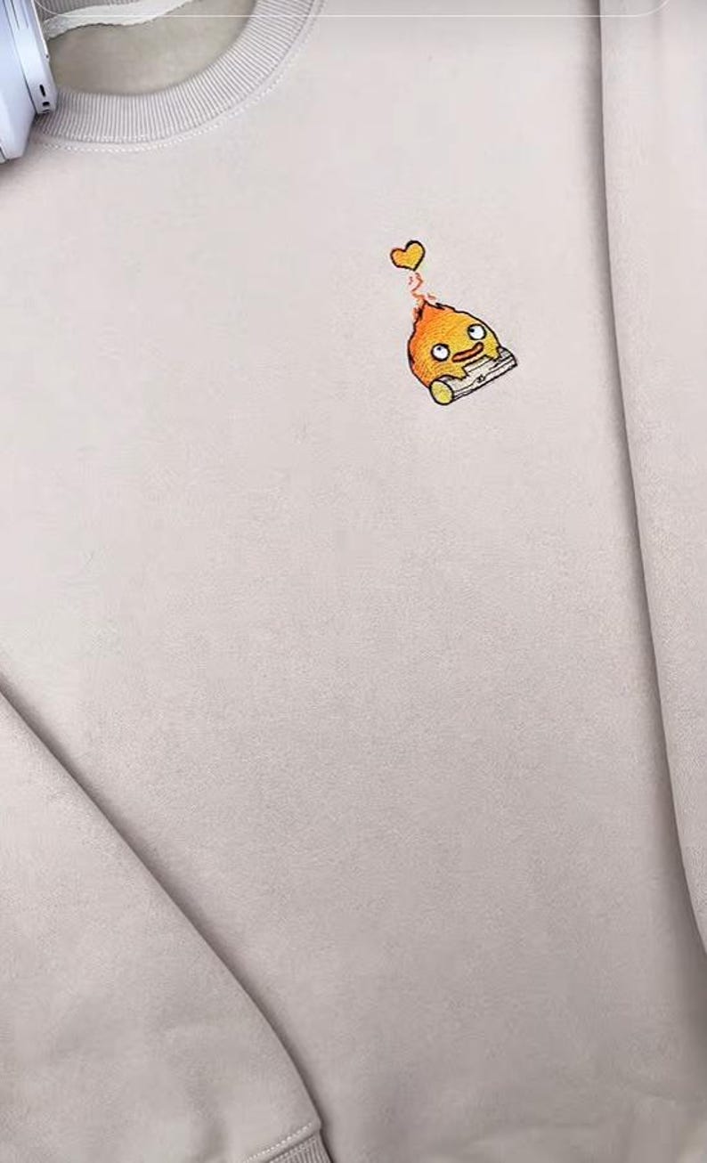 Op de afbeelding: Een lichtbeige sweatshirt met een klein geborduurd cartoonfiguur. Het figuur is oranje met een gele vlam en een klein hartje erboven. De sweatshirt heeft een ronde hals en lange mouwen.