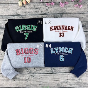 May include: Four folded sweatshirts with text. The top left is black with "GIBSIE" in green, "Boys of Tommen", and the number 7. The top right is white with "KAVANAGH", "Boys of Tommen", and the number 13. The bottom left is grey with "BIGGS", "Boys of Tommen", and the number 10. The bottom right is navy blue with "LYNCH", "Boys of Tommen", and the number 6.