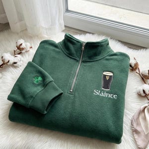 May include: A dark green quarter-zip sweatshirt with a silver zipper. The sweatshirt features a shamrock on the left cuff and an embroidered pint of stout with the word "Sl&aacute;inte" in white. The sweatshirt is on a white fluffy surface.