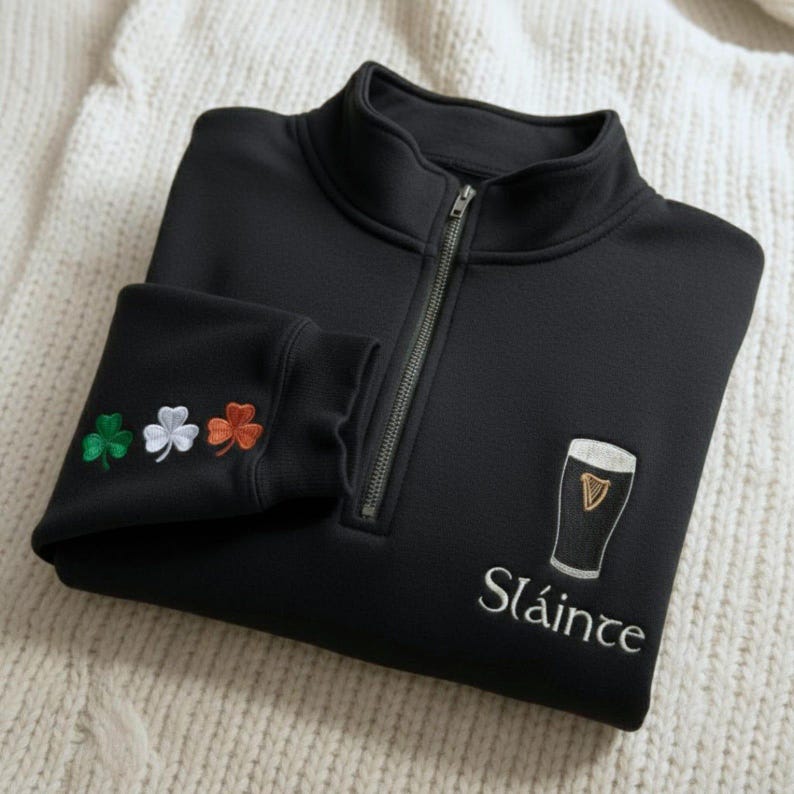 May include: Black quarter-zip sweatshirt with a white embroidered pint of stout and the word "Sl&aacute;inte". The left sleeve cuff features embroidered shamrocks in green, white, and orange. The sweatshirt is folded on a white, textured surface.