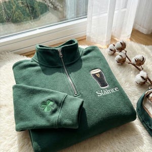May include: A green quarter-zip sweatshirt with a high collar and a silver zipper. The sweatshirt features an embroidered shamrock on the left sleeve and a graphic of a pint of stout with the word "Sl&aacute;inte" below it.