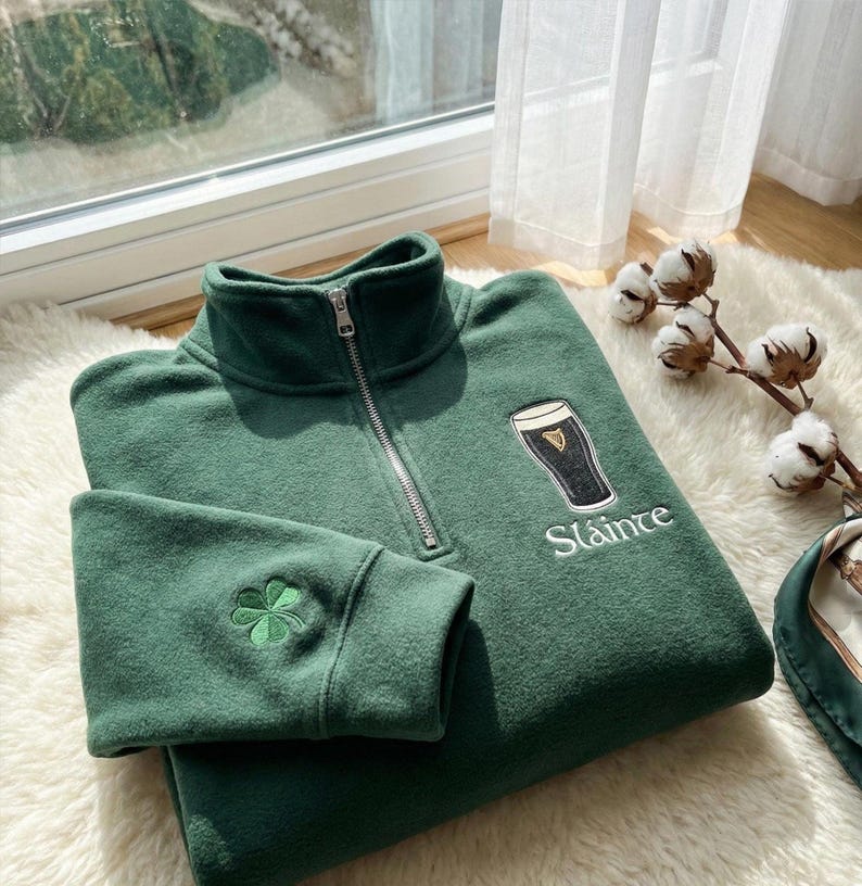 May include: A green quarter-zip sweatshirt with a high collar and a silver zipper. The sweatshirt features an embroidered shamrock on the left sleeve and a graphic of a pint of stout with the word "Sl&aacute;inte" below it.