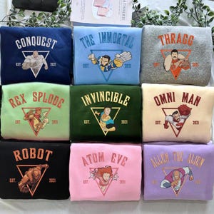 Könnte beinhalten: Eine Sammlung von neun Sweatshirts mit Rundhalsausschnitt in verschiedenen Farben, jeweils mit einem einzigartigen Grafikdesign und Text. Die Designs umfassen Charaktere und Texte wie "CONQUEST", "THE IMMORTAL" und "ROBOT". Auf jedem Sweatshirt steht "EST. 2021".