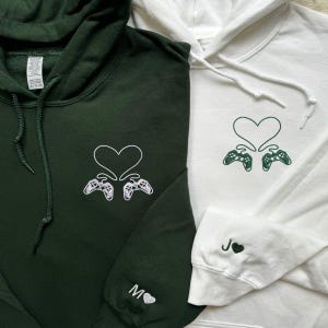 May include: Two hoodies, one dark green and one white. Each features a heart and two game controllers embroidered on the front. The dark green hoodie has an "M" and a heart on the sleeve, the white one has a "J" and a heart.