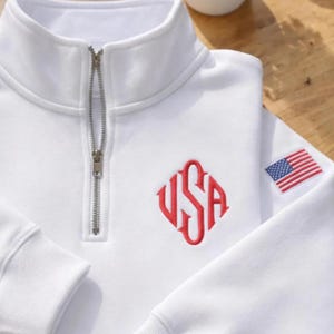 May include: White quarter-zip sweatshirt with a red "USA" monogram and an American flag patch on the sleeve. The sweatshirt has a silver zipper and a high collar. The fabric appears to be a soft, comfortable material.