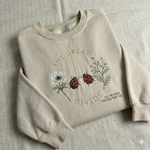 May include: Beige sweatshirt with embroidered design. The design includes the text "THE GREAT DIVIDE" and "CALL ME WHEN THE BUGS DON'T DIE". The design also features two red ladybirds, white flowers, and green stems.