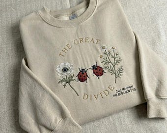 May include: Beige sweatshirt with embroidered design. The design includes the text "THE GREAT DIVIDE" and "CALL ME WHEN THE BUGS DON'T DIE". The design also features two red ladybugs, white flowers, and green stems.