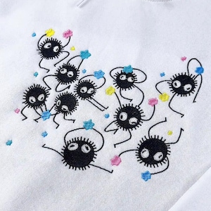 May include: White hoodie with an embroidered design of black, round, cartoon-like creatures with white eyes and thin limbs. The design includes colourful star-shaped accents in pink, yellow, and blue.
