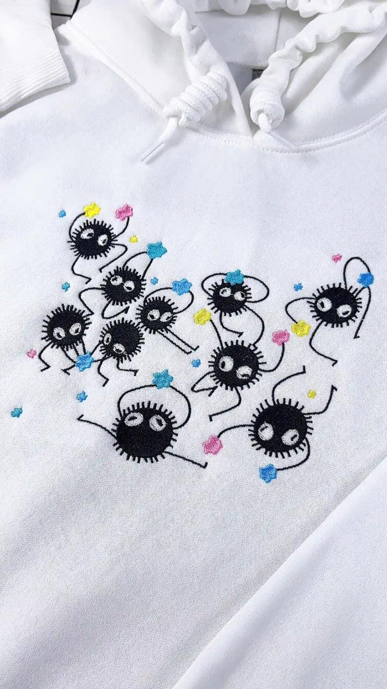 May include: White hoodie with an embroidered design of black, round, cartoon-like creatures with white eyes and thin limbs. The design includes colourful star-shaped accents in pink, yellow, and blue.