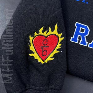 May include: Black sweatshirt sleeve with a red heart-shaped emblem, outlined in yellow flames. The heart contains the letters "c/b" in black. The word "MHFulfillment" is printed vertically on the left side of the sleeve.