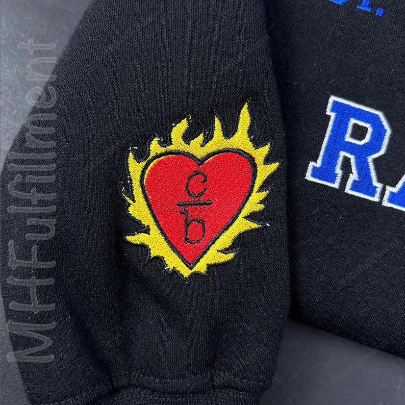 May include: Black sweatshirt sleeve with a red heart-shaped emblem, outlined in yellow flames. The heart contains the letters "c/b" in black. The word "MHFulfillment" is printed vertically on the left side of the sleeve.
