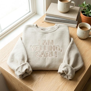May include: A beige sweatshirt with the embroidered text "I AM YELLING, CARL!". The sleeves are cuffed. Two mugs of coffee and a small potted plant sit on a wooden table next to the sweatshirt.