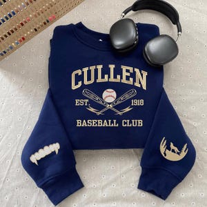 May include: Navy blue sweatshirt with gold embroidered text reading "CULLEN BASEBALL CLUB EST. 1918". The sleeves have gold embroidered designs of fangs and a wolf in a forest. Black headphones are placed on the sweatshirt.