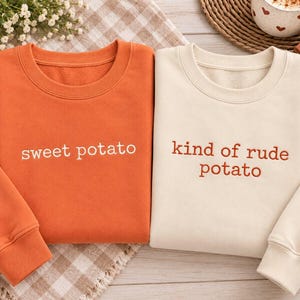 May include: Two crewneck sweatshirts. The orange sweatshirt features the white embroidered text "sweet potato". The cream-colored sweatshirt features the orange embroidered text "kind of rude potato". Both have long sleeves and ribbed cuffs.