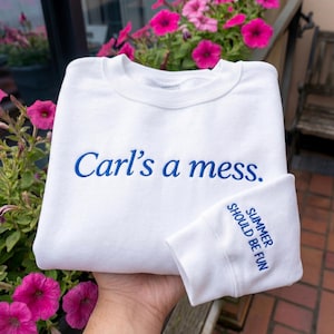 May include: A white crewneck sweatshirt featuring blue embroidery on the chest that reads Carl's a mess. The sleeve cuff also features blue embroidery that reads SUMMER SHOULD BE FUN. The garment has a ribbed collar and long sleeves.
