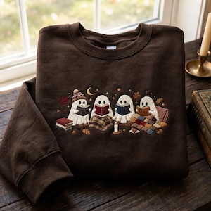 Puede incluir: Una sudadera de cuello redondo color marrón oscuro con un diseño bordado de cuatro fantasmas blancos leyendo libros. Los fantasmas están rodeados de pequeñas hojas de otoño, una luna creciente, estrellas, una vela encendida y libros.