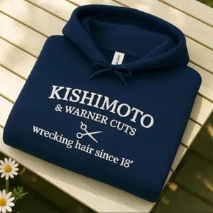 May include: A navy blue hooded sweatshirt with white embroidered text that reads "KISHIMOTO & WARNER CUTS wrecking hair since 18'" and a pair of scissors graphic. The sweatshirt has a drawstring hood.