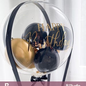 May include: A clear balloon with black and gold balloons inside, and the words "Happy Birthday" in gold script. The balloon is decorated with black ribbon and a black bow at the base. The balloon is a festive decoration.