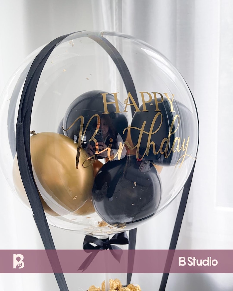 May include: A clear balloon with black and gold balloons inside, and the words "Happy Birthday" in gold script. The balloon is decorated with black ribbon and a black bow at the base. The balloon is a festive decoration.