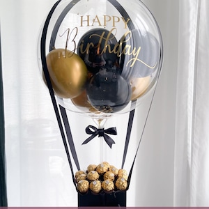 May include: A clear balloon with "Happy Birthday" in gold script, containing gold and black balloons. The balloon is suspended above a black box filled with gold-wrapped chocolates, with black ribbons and a bow.