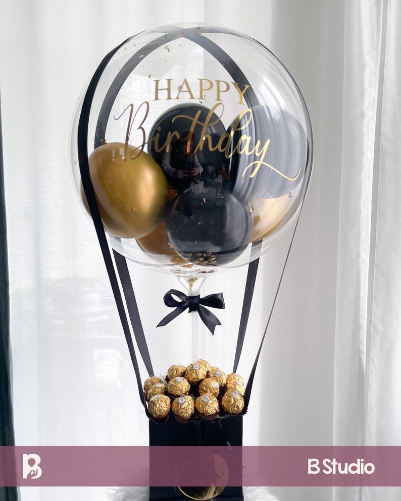 May include: A clear balloon with "Happy Birthday" in gold script, containing gold and black balloons. The balloon is suspended above a black box filled with gold-wrapped chocolates, with black ribbons and a bow.
