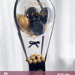 May include: A clear balloon with gold and black balloons inside, and the words "Happy Birthday" in gold script. The balloon is attached to a black box filled with gold-wrapped chocolates, and a black bow.