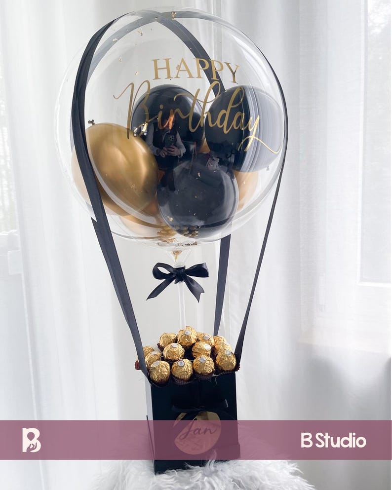 May include: A clear balloon with gold and black balloons inside, and the words "Happy Birthday" in gold script. The balloon is attached to a black box filled with gold-wrapped chocolates, and a black bow.