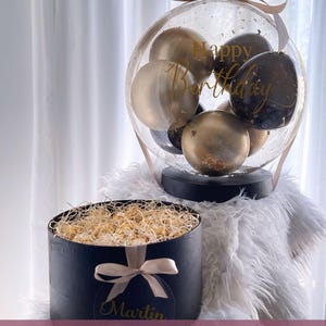 May include: A black gift box with a gold ribbon and the name "Martin" on it. The box is filled with gold-wrapped chocolates. A clear balloon with black and gold balloons inside is decorated with the text "Happy Birthday".