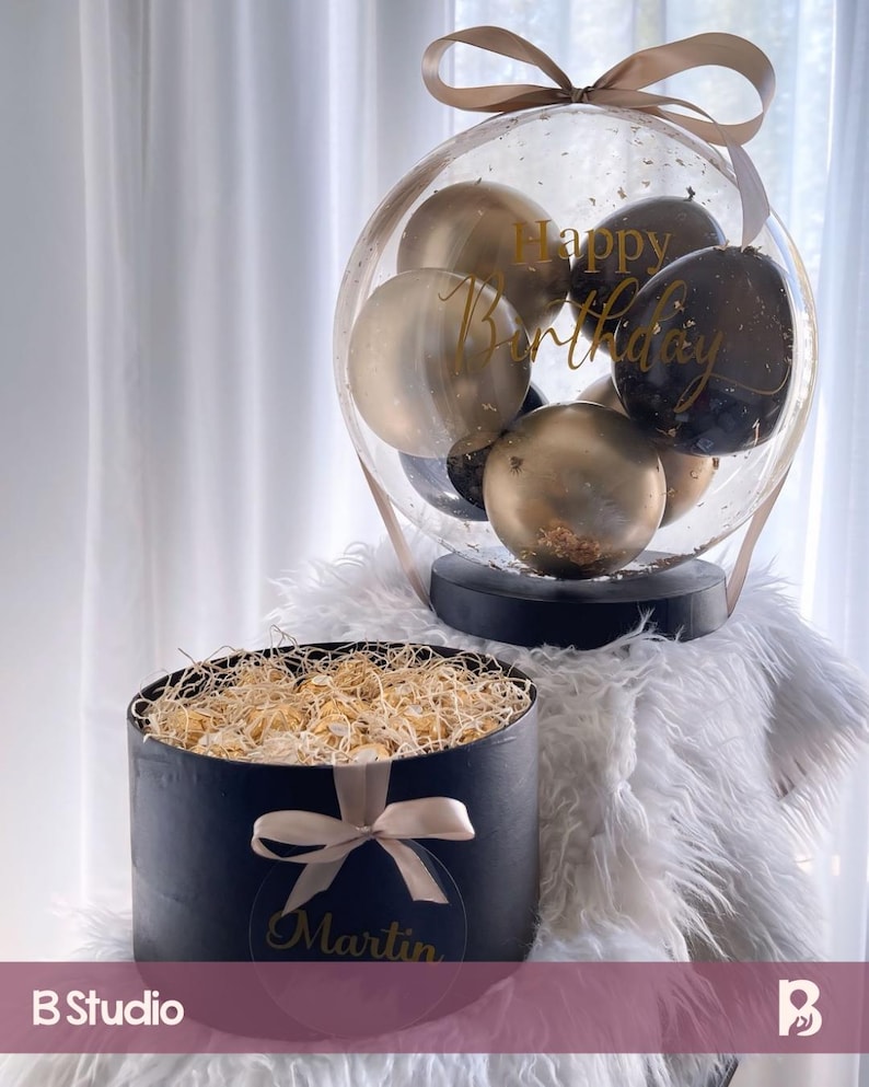 May include: A black gift box with a gold ribbon and the name "Martin" on it. The box is filled with gold-wrapped chocolates. A clear balloon with black and gold balloons inside is decorated with the text "Happy Birthday".