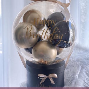 May include: A clear balloon filled with gold and black balloons. The balloon is tied with a beige ribbon and sits on top of a black box with a beige ribbon and a gold name tag. The balloon has the words "Happy Birthday" written on it.