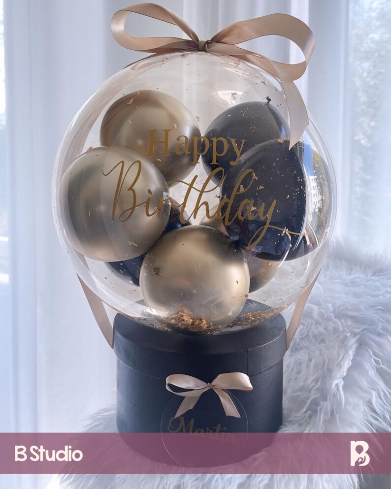 May include: A clear balloon filled with gold and black balloons. The balloon is tied with a beige ribbon and sits on top of a black box with a beige ribbon and a gold name tag. The balloon has the words "Happy Birthday" written on it.
