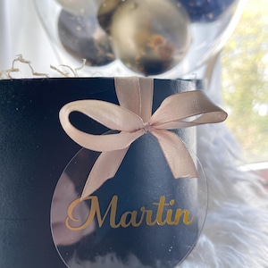May include: A black gift box with a beige ribbon and a clear round tag with the name "Martin" written in gold lettering.
