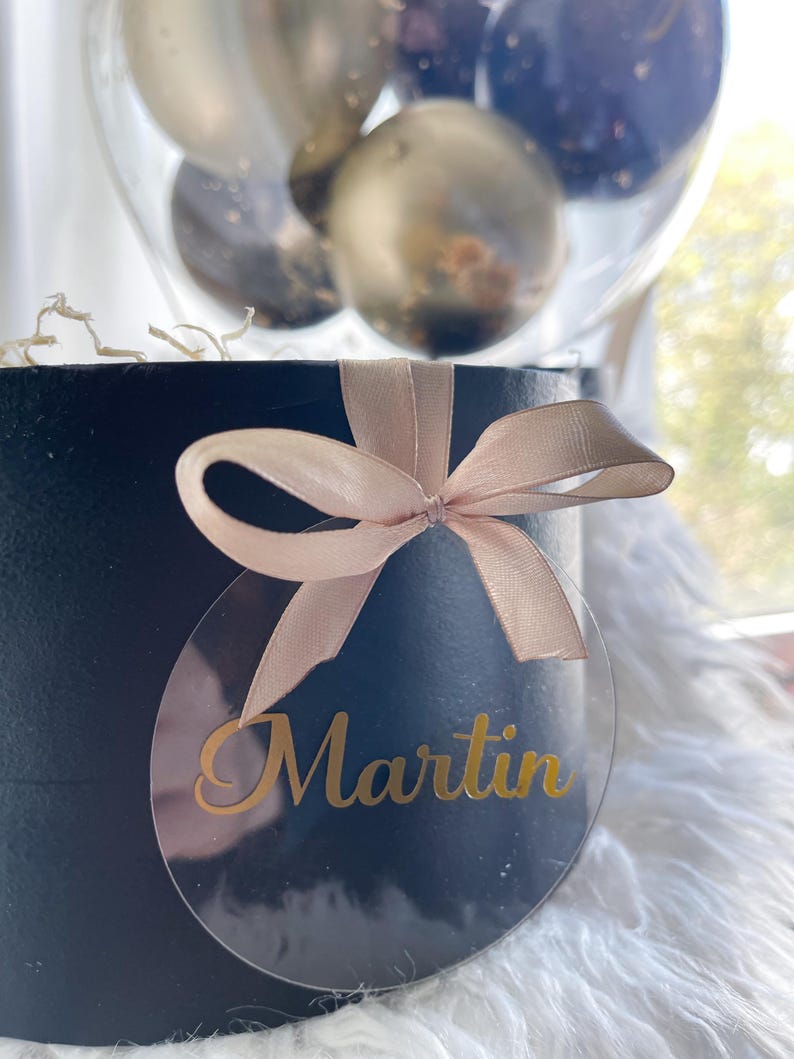 May include: A black gift box with a beige ribbon and a clear round tag with the name "Martin" written in gold lettering.