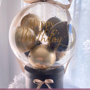 May include: A clear balloon filled with black and gold balloons, with the words "Happy Birthday" written on it. The balloon is attached to a black box with a gold ribbon and a clear circle with the name "Martin" written on it.
