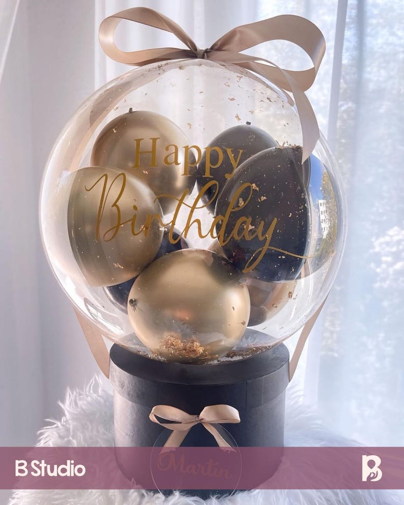 May include: A clear balloon filled with black and gold balloons, with the words "Happy Birthday" written on it. The balloon is attached to a black box with a gold ribbon and a clear circle with the name "Martin" written on it.