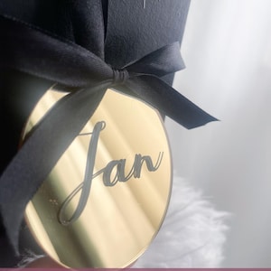 May include: A gold-colored, circular tag with the name "Jan" in cursive script, attached to a black ribbon tied in a bow. The tag is hanging from a black gift box. The background is a soft, white, feathery texture.