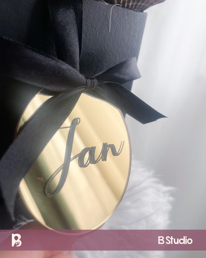 May include: A gold-colored, circular tag with the name "Jan" in cursive script, attached to a black ribbon tied in a bow. The tag is hanging from a black gift box. The background is a soft, white, feathery texture.