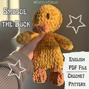 May include: A handmade, plush yellow duck toy with orange feet and beak. The duck has a black eye and is labeled "Smudge the Duck." The image also includes text that says "English PDF File Crochet Pattern."