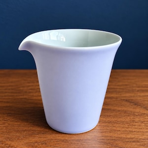May include: A light lavender ceramic pitcher with a smooth, matte finish and a flared rim featuring a small, integrated pouring spout. The vessel has a tapered body that narrows toward the base, showcasing a clean, simple design.