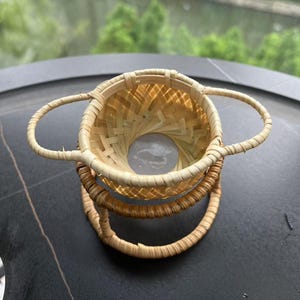 May include: A small, woven bamboo basket with two handles. The basket has a circular base and a flared top. The basket is made of light brown bamboo and has a natural, rustic appearance. The basket is sitting on a dark surface.