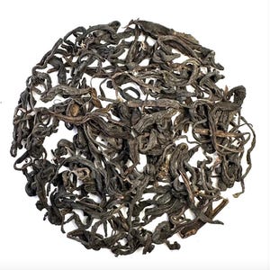 May include: A close-up view of a circular arrangement of dark, dried tea leaves against a white background. The leaves are a deep brown color with a slightly curled and twisted appearance, suggesting a high-quality tea product.