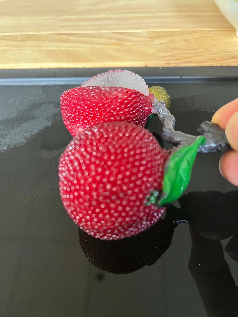 以下が含まれることがあります： 赤い質感のあるベリーのようなオブジェクトのクローズアップ。茎と緑の葉が付いています。オブジェクトはでこぼこの表面を持ち、暗い背景に配置されています。茎はグレーで、葉は鮮やかな緑色です。