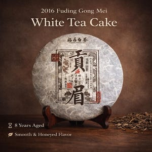 May include: A round, aged white tea cake from 2016, displayed on a wooden stand. The tea cake has a textured, light gray surface with a rectangular label featuring Chinese characters. The text on the image reads "2016 Fuding Gong Mei White Tea Cake".