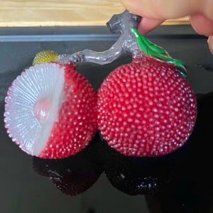 以下が含まれることがあります： 赤いライチの実2つのクローズアップ。1つはカットされ、白い果肉と種が見える。実はざらざらした表面で、緑の葉が付いた灰色の茎に付いている。ライチは手で持たれている。